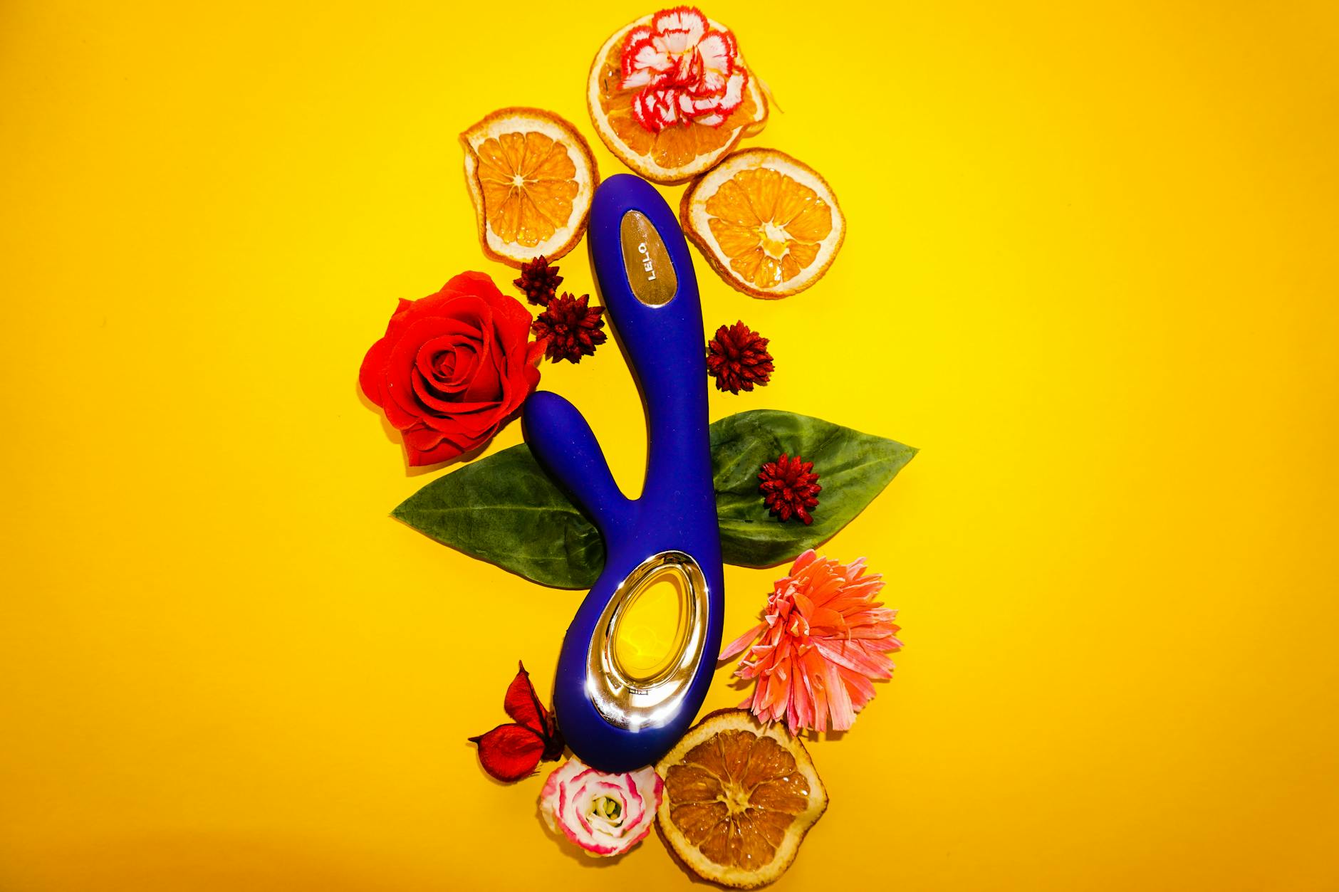 A lemon-colored vibrator surrounded by flowers and citrus on a bright yellow surface
